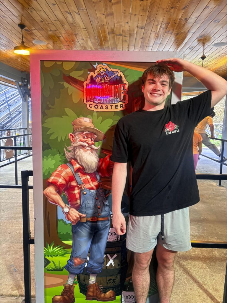 Standing next to a Moonshine Mountain Coaster height chart featuring a cartoon moonshiner and a barrel.