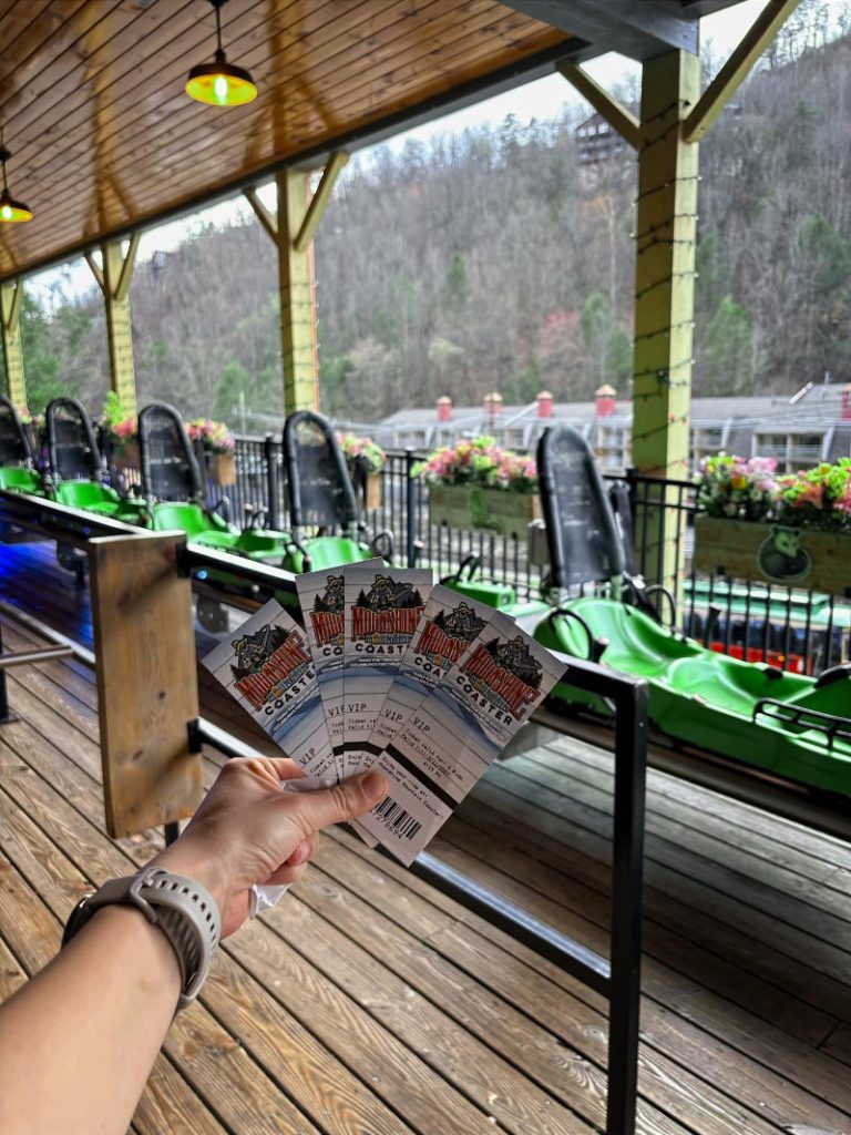 hand holding four VIP tickets in front of green mountain coaster cars lined up at the loading station.