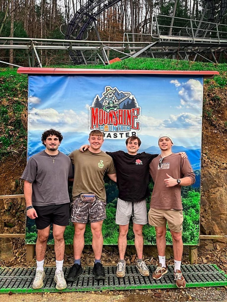 Calvin Orley and three friends standing in front of a Moonshine Mountain Coaster backdrop with the coaster track visible in the background.