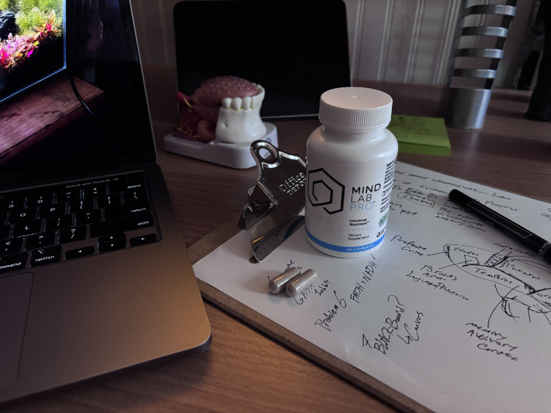 A detailed photograph of a clean, organized two-tiered wooden study desk, featuring a bottle of MIND LAB PRO V4 Universal Nootropic and open capsules. Handwritten logic notes, a tablet displaying a muscle anatomy model, a terrarium, a dental model, and a laptop showing an Ohio University course portal for a pre-med Philosophy class are arranged on the desk. This is where Calvin optimizes his cognitive focus while studying at OU.